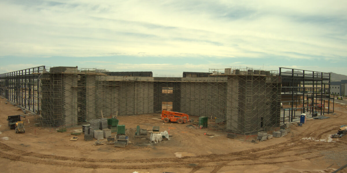 Construction site showing a large building framework with scaffolding, construction materials, and equipment scattered on dirt ground under a cloudy sky.
