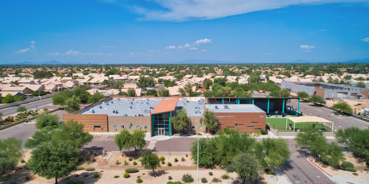 Aerial view of a modern school building surrounded by trees and residential houses under a clear blue sky.