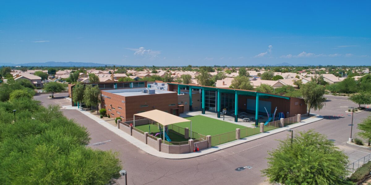 Aerial view of a modern brick school building with a covered outdoor area and artificial turf, surrounded by suburban houses and trees under a clear blue sky.