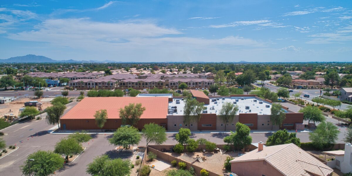 Aerial view of a large, flat-roofed building surrounded by trees and parking lots in a suburban neighborhood under a blue sky.