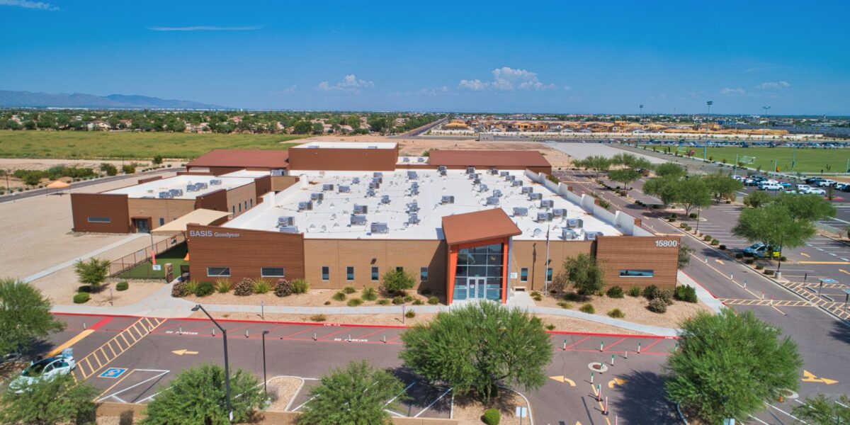 Aerial view of a modern school building labeled "BASIS Goodyear," surrounded by parking lots, trees, and desert landscape under a clear blue sky.