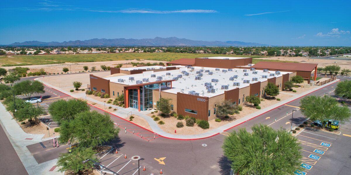 Aerial view of a modern, single-story commercial or institutional building with a large parking lot and surrounding desert landscaping.