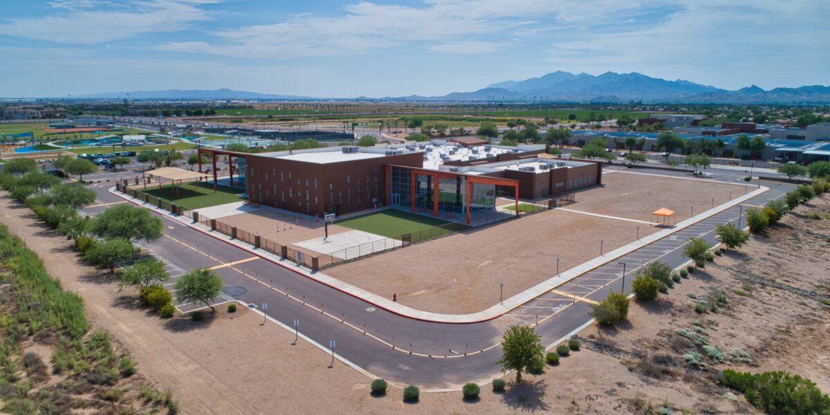 A modern school building with outdoor play areas, surrounded by a desert landscape and mountains in the distance, under a partly cloudy sky.