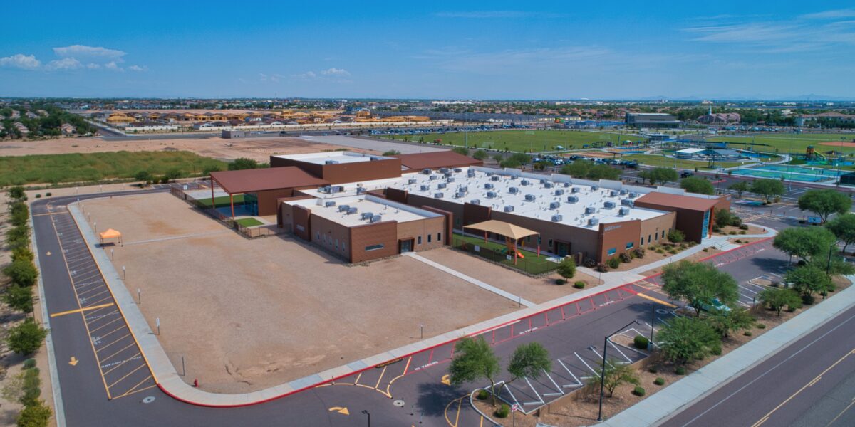 Aerial view of a modern school building with white roofs, surrounding parking lots, and adjacent open fields under a clear blue sky.