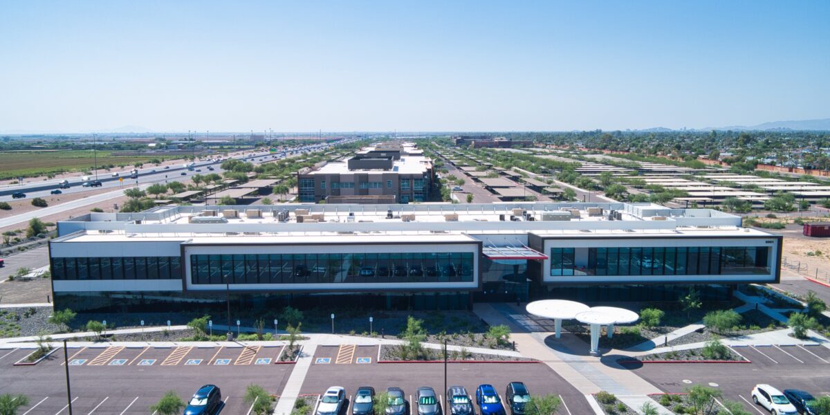 Aerial view of a modern, single-story building with large windows, a parking lot with cars, and a highway and fields in the background under a clear sky.