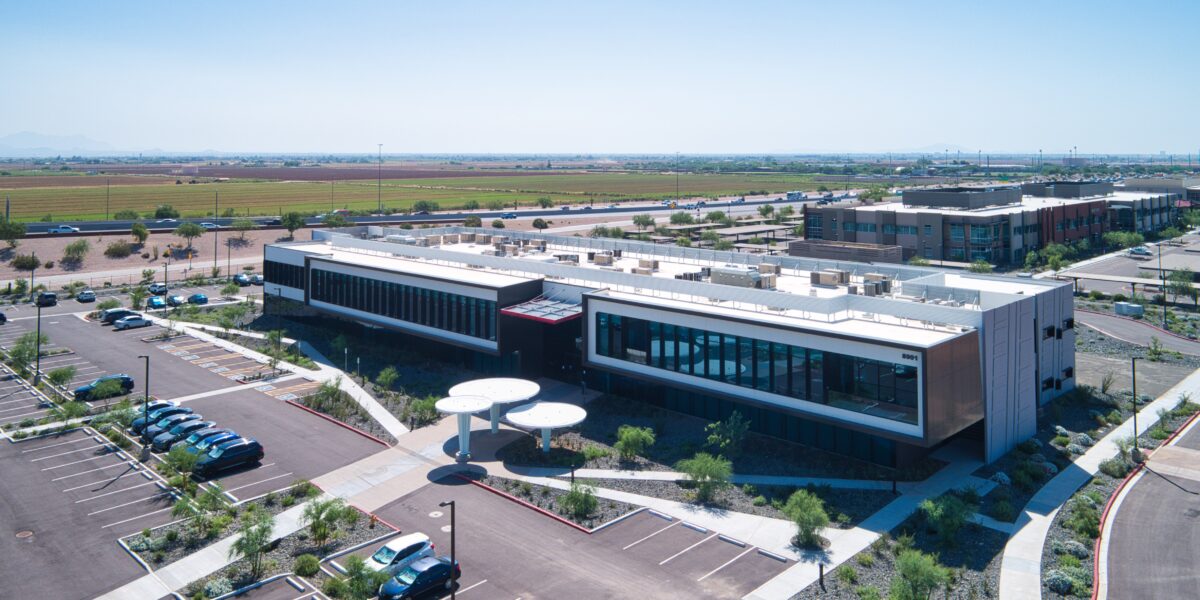 Aerial view of a modern, rectangular commercial building with large windows, surrounded by parking lots, landscaping, and adjacent buildings under a clear sky.