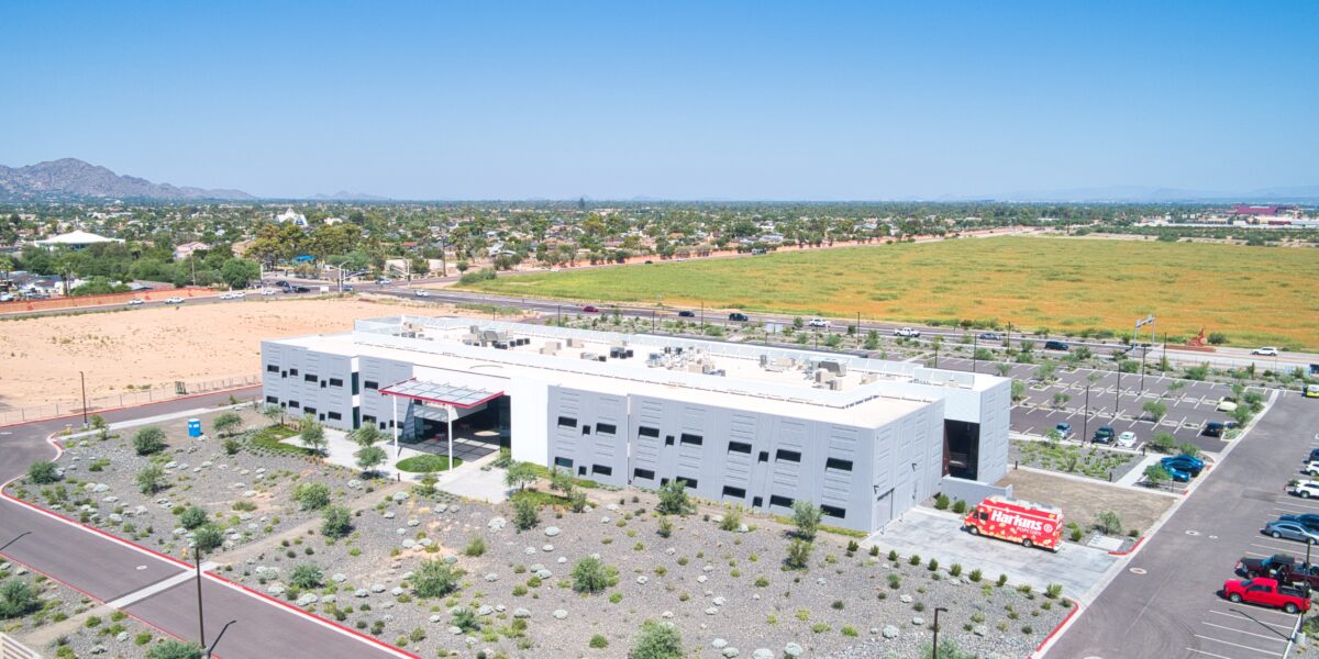 Aerial view of a large, modern, white commercial building surrounded by parking lots and desert landscaping, with open fields and distant mountains in the background.
