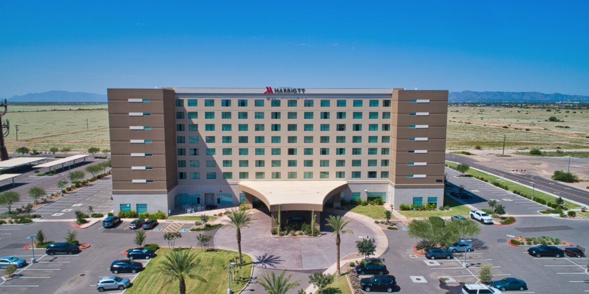 A six-story Marriott hotel with a parking lot in front, surrounded by desert landscape and mountains in the distance under a clear blue sky.