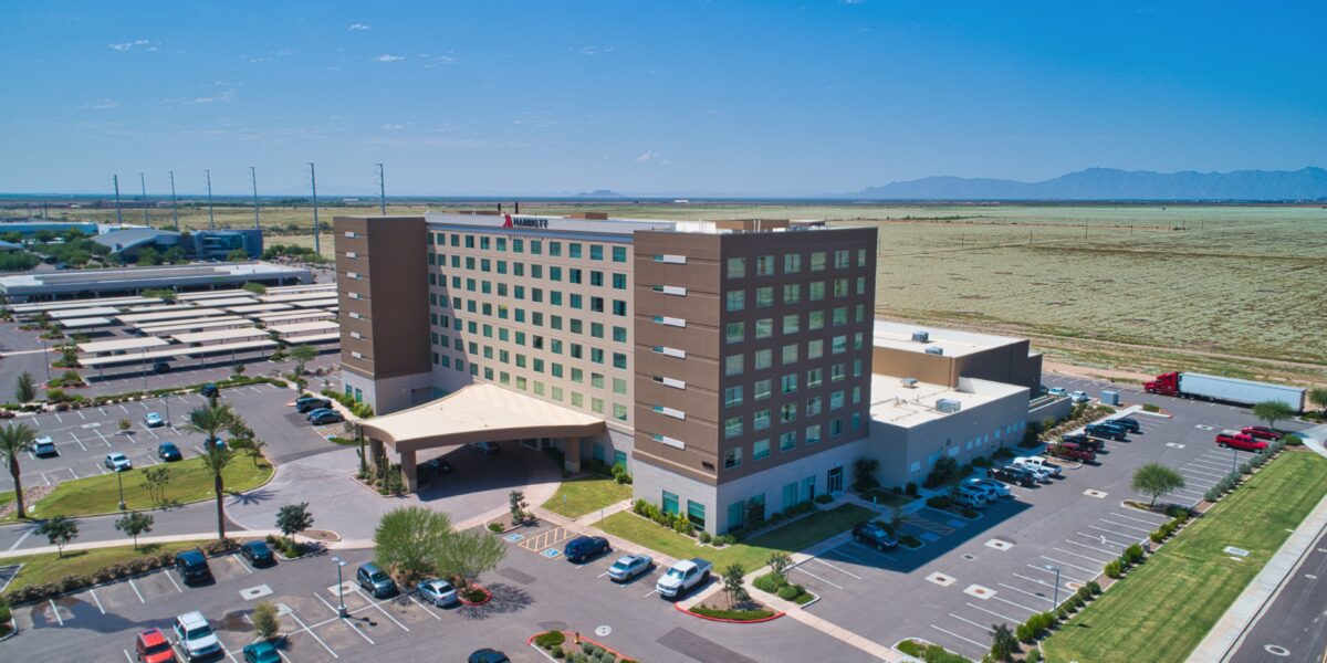 A large, multi-story hospital building with a covered entrance, surrounded by parking lots and open fields under a clear blue sky.