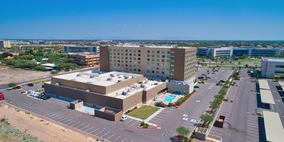 Aerial view of a mid-rise hotel building with a pool, surrounded by parking lots and office buildings under a clear sky.