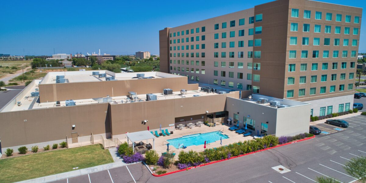 A mid-rise hotel building with a rectangular outdoor pool, lounge chairs, and umbrellas. There are parking spaces and landscaped areas in the foreground under a clear blue sky.