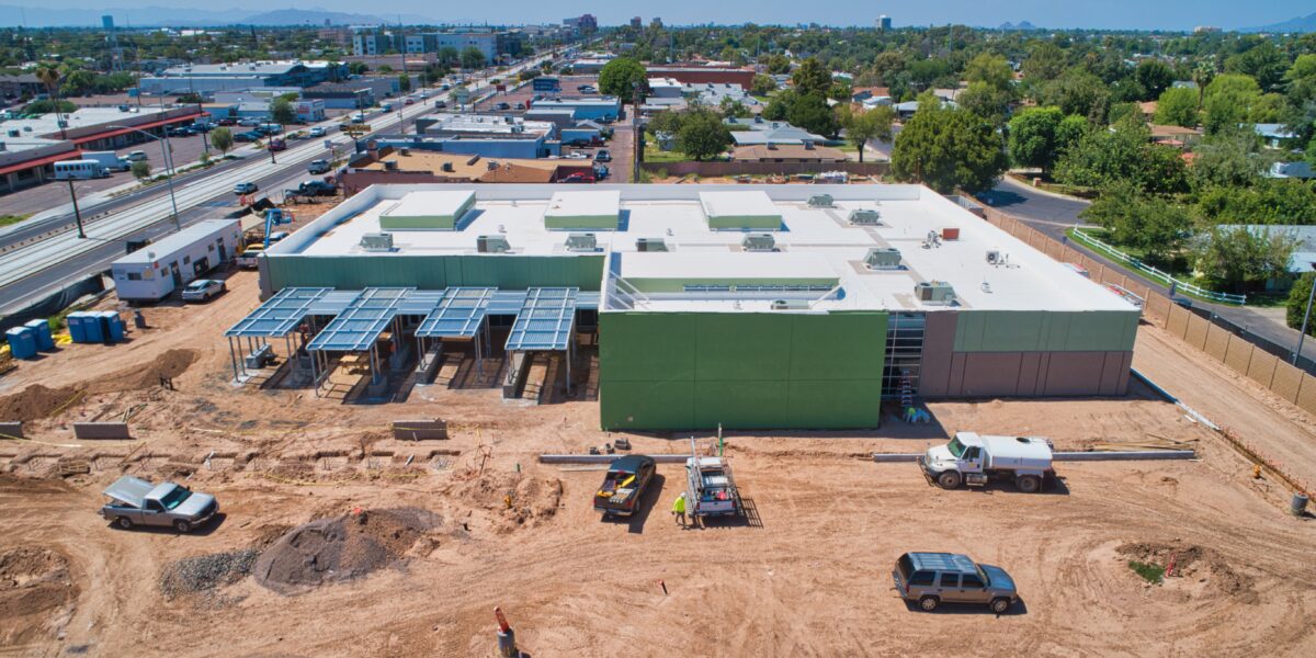 Aerial view of a construction site with a large green building, solar panel structures, vehicles, and equipment on a dirt lot in an urban area.