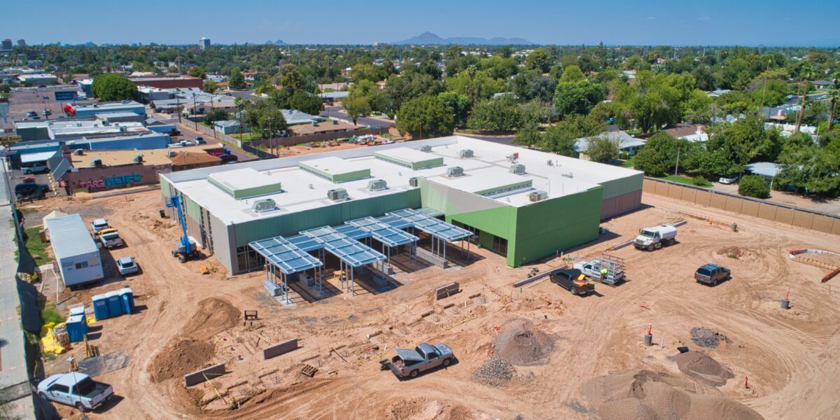 Aerial view of a construction site featuring a large, modern building with solar panel structures in a suburban area, surrounded by vehicles and construction materials.
