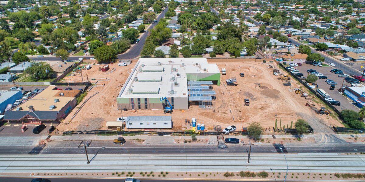 Aerial view of a large building under construction surrounded by dirt, vehicles, and a residential neighborhood. A main road and light rail tracks run in front of the site.
