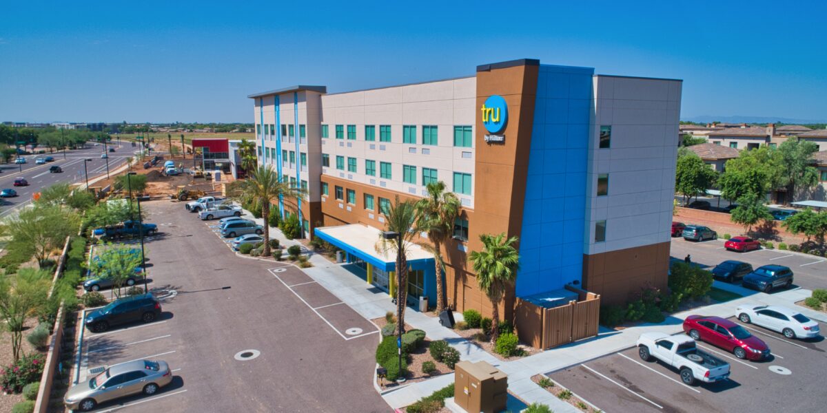 Aerial view of a modern four-story Tru by Hilton hotel building with a parking lot, palm trees, and nearby roads under clear blue sky.