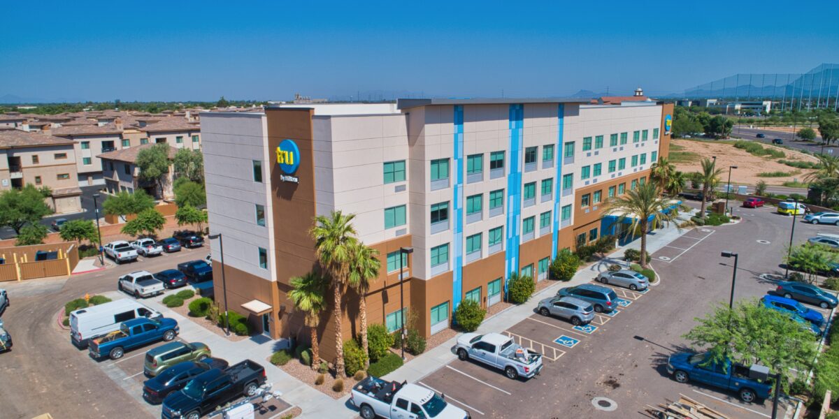 A four-story hotel building with blue accents, a parking lot with cars, palm trees, and neighboring buildings under a clear blue sky.