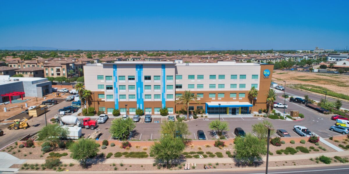 A four-story hotel building with blue accents, a parking lot in front, and landscaped surroundings, set in a suburban area with houses and open land nearby.