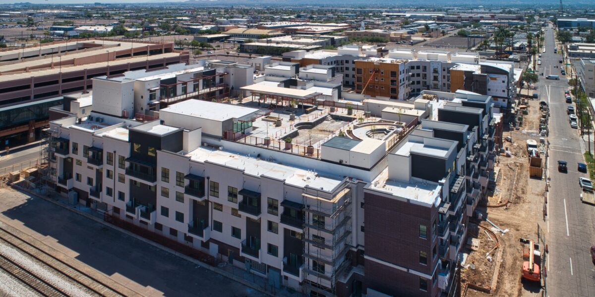 Aerial view of a large, modern apartment complex under construction in an urban area, with nearby roads, industrial buildings, and mountains in the distance.