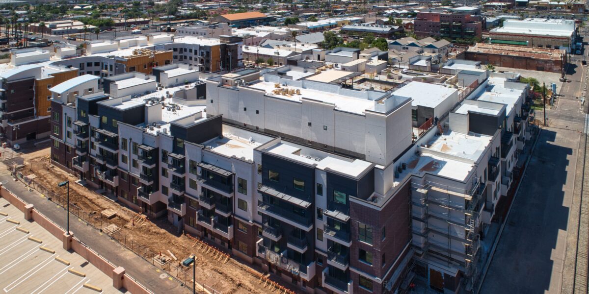 Aerial view of a large, modern apartment complex under construction in an urban area, with nearby buildings, parking lot, and distant mountains visible.