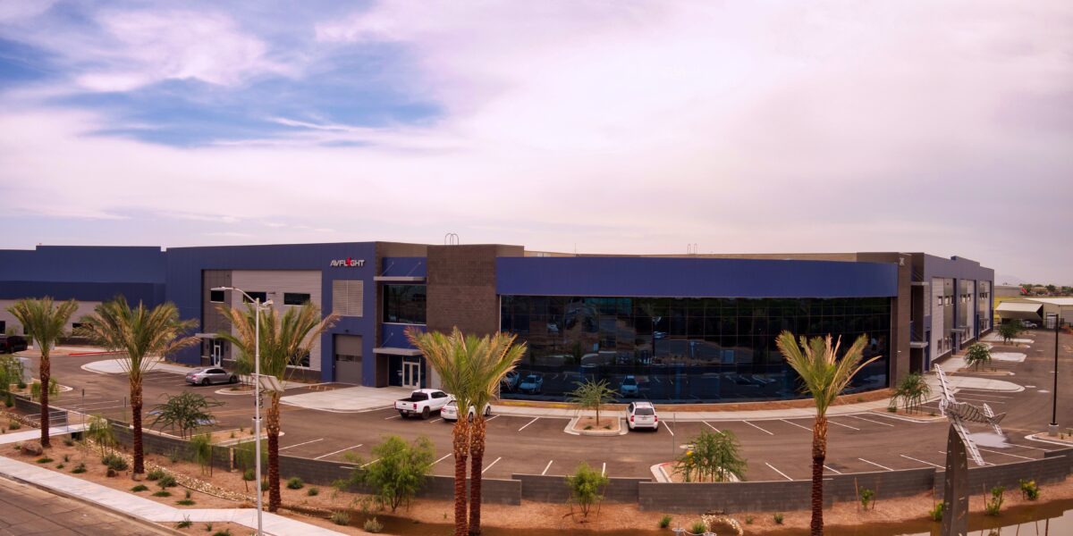 Modern commercial building with large glass windows, palm trees, and several parked vehicles in a mostly empty parking lot under a partly cloudy sky.